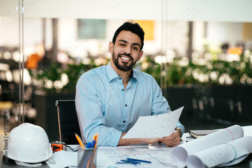 Happy attractive millennial arabic engineer with beard works with documents at workplace with hard hat