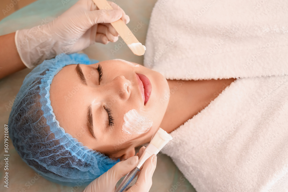 Cosmetologist applying mask onto young woman's face in beauty salon