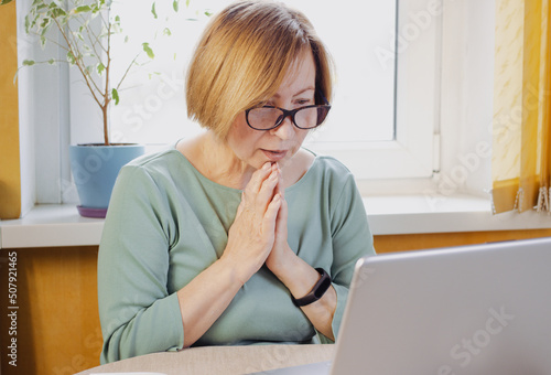 Socked surprised mature woman in eyeglasses looking at laptop reading disturbing news in media, watching video content, sitting indoors. Feeling hope, praying for happy end. Received bad email
