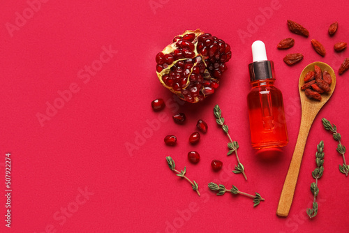 Bottle of natural serum, spoon with goji berries, thyme and pomegranate on red background