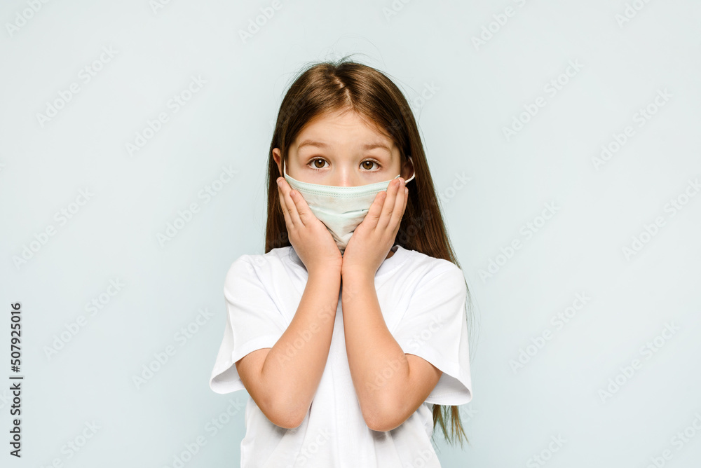 Frightened caucasian little girl is feeling surprised in a medical mask on a blue background. Copy space. Protection against viruses, coronavirus and pandemic. Frightened caucasian little girl is feeling surprised in a medical mask on a blue background. Copy space. Protection against viruses, coronavirus and pandemic.
