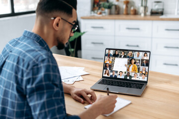 Distance learning, online course. View over the shoulder of a guy taking notes in a notebook, at a laptop screen with a teacher and a group of multiracial students. E-learning, webinar, online lecture