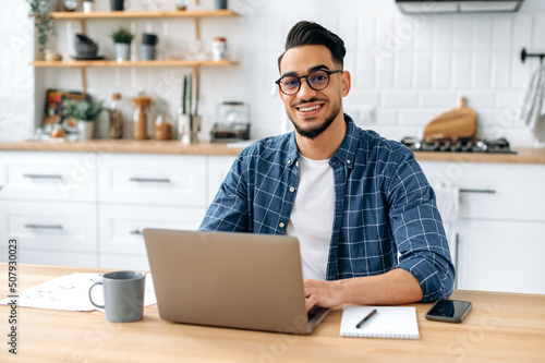 Portrait of a handsome successful confident Arabian or Indian guy with glasses  freelancer  designer or IT specialist  sitting at home in the kitchen  working online by laptop  looks at camera  smiles