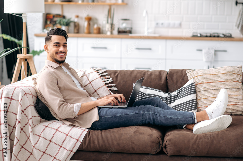 Attractive smiling arabian or indian man in casual stylish wear, freelancer or student, sitting on couch in living room, working on a project uses laptop, looking at the camera, smiling happily Attractive smiling arabian or indian man in casual stylish wear, freelancer or student, sitting on couch in living room, working on a project uses laptop, looking at the camera, smiling happily