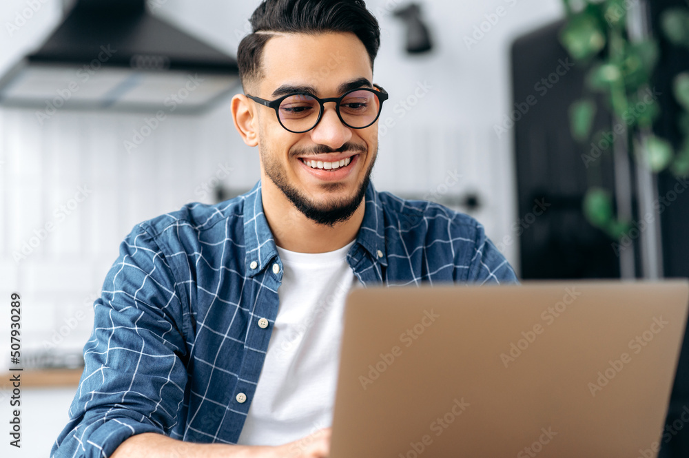 Close-up photo of attractive positive indian or arabian guy with glasses, freelancer or creative designer, sitting at a desk at home, working on a creative project uses laptop, looks at screen, smiles Close-up photo of attractive positive indian or arabian guy with glasses, freelancer or creative designer, sitting at a desk at home, working on a creative project uses laptop, looks at screen, smiles