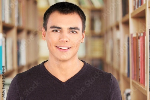 Happy schoolboy guy posing in campus library on bookshelves background.