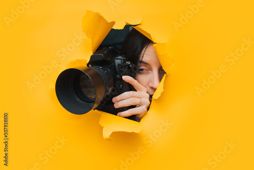 A young caucasian paparazzi girl holds a reflex camera and looks through a torn hole in yellow paper. The concept of embarrassment, shame for what he saw. Copy space.