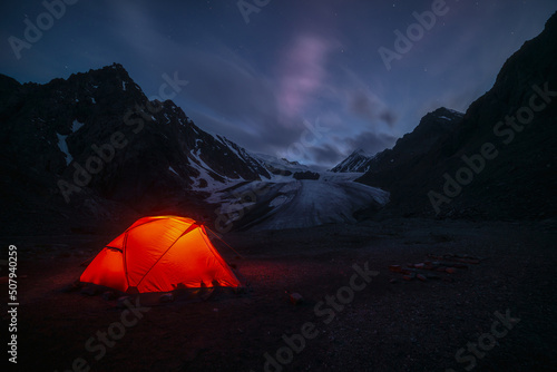 Awesome mountain landscape with vivid orange tent near large glacier tongue under clouds in night starry sky. Tent glow by orange light with view to glacier and mountains silhouettes in starry night.