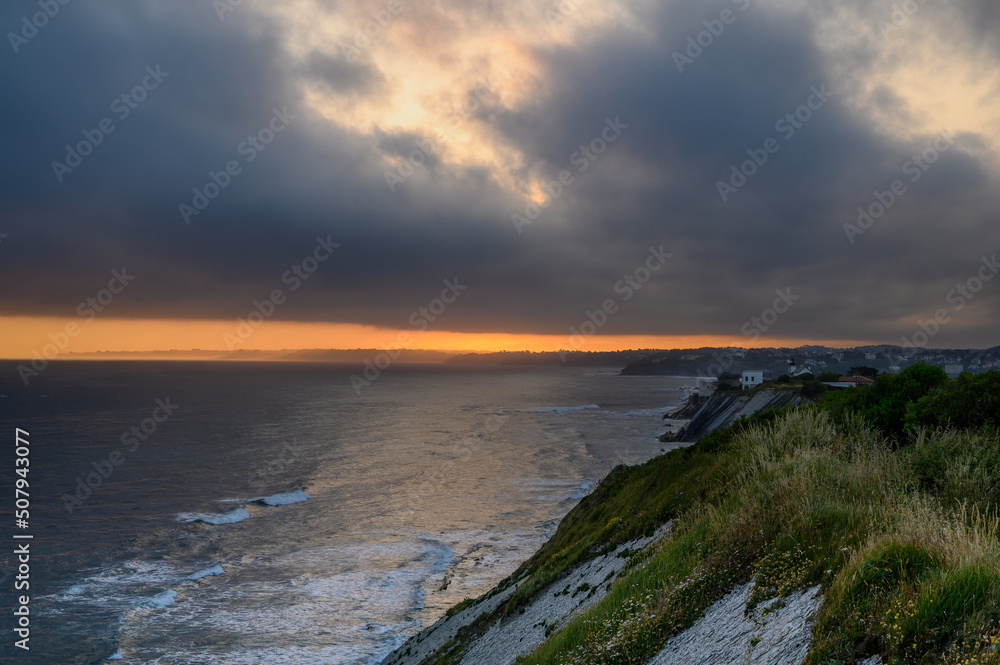 View of the French Basque coast at dawn on a spring day. Below the dark sea on the right side the coast in front a strip of orange color lit like fire that is the coastline, in the sky a large dark gr