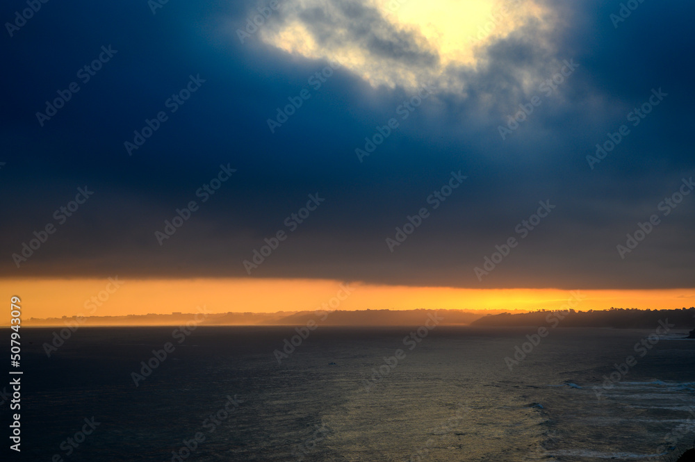 View of the French Basque coast at dawn on a spring day. Below the dark sea on the right side the coast in front a strip of orange color lit like fire that is the coastline, in the sky a large dark gr