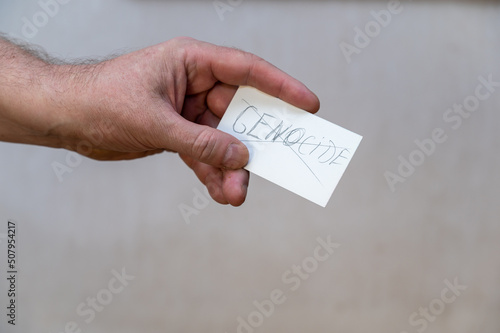 The word GENOCIDE is crossed out. A word written in jagged letters. A man's hand holds a white paper rectangle with text against a gray background photo