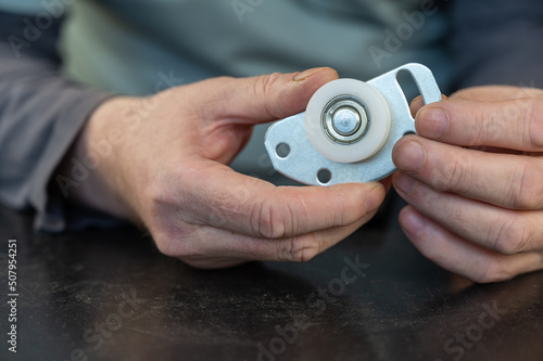 A grown man holds in his lower roller for a sliding door. Metal mounting plate with white plastic roller. Furniture hardware. Close-up. Selective focus.