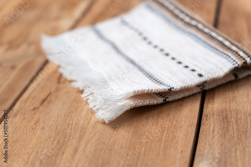 A white kitchen towel lies on the countertop of a wooden table. The towel is fringed and patterned on the rough boards. Selective Focus.