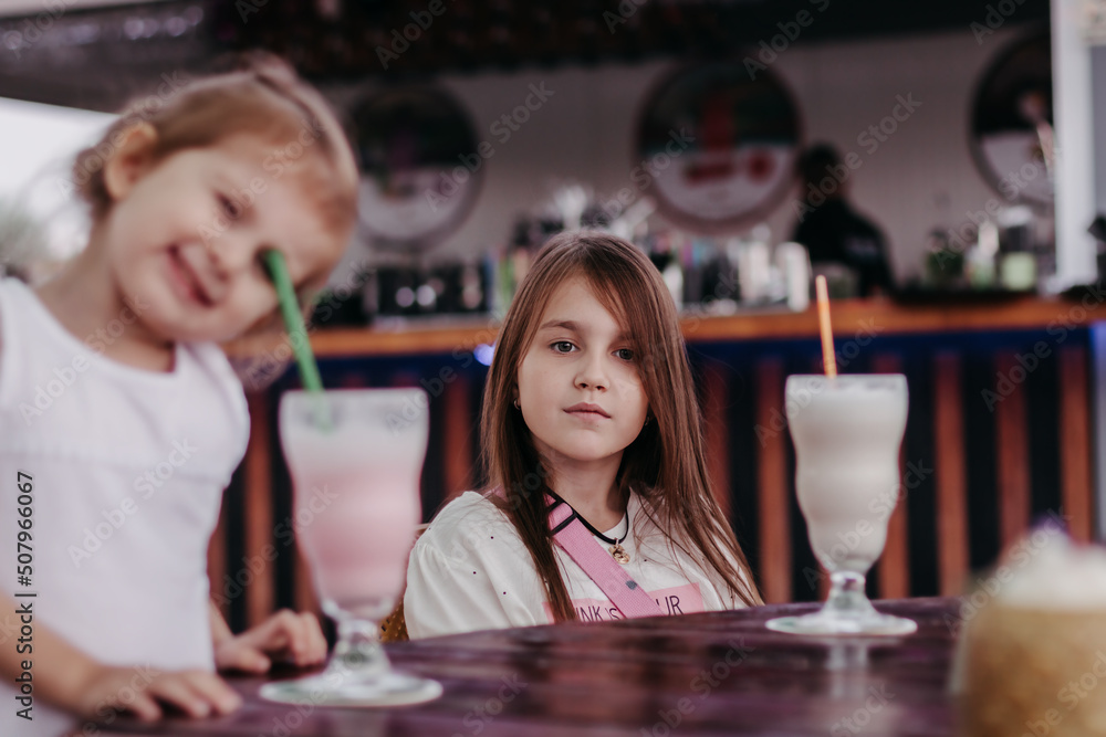 Cute little girl wearing white shirt sitting at table in cafe drinking milk shake, lifestyle portrait
