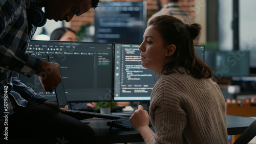 Junior programer sitting down on desk showing laptop with source code to senior dev asking for opinion. Software developer writing algorithm interrupted by colleague wanting help with fixing errors.