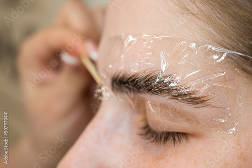 The master applies the composition for laminating the eyebrows to the client with a wooden spatula. 