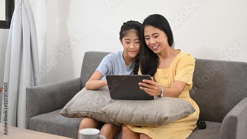 Smiling Asian mother and daughter using digital tablet while sitting on sofa, enjoying spending free time together at home