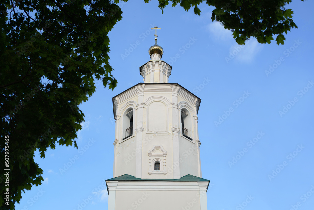 Close-up of white church with cross against blue sky, an old monastery in frame of trees outdoors