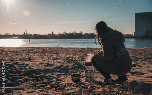 Young asian woman with warm clothes on is cooking with outdoor cooker on Harriersand beach at a riverbank during golden hour. Weser river blurred in the background. 