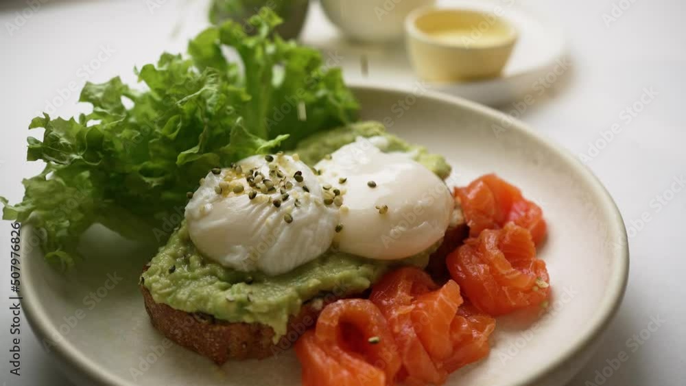 Chef sprinkles seeds on toast with poached eggs and guacamole. Delicious toast served with trout slices and fresh salad leaves extreme closeup Chef sprinkles seeds on toast with poached eggs and guacamole. Delicious toast served with trout slices and fresh salad leaves extreme closeup
