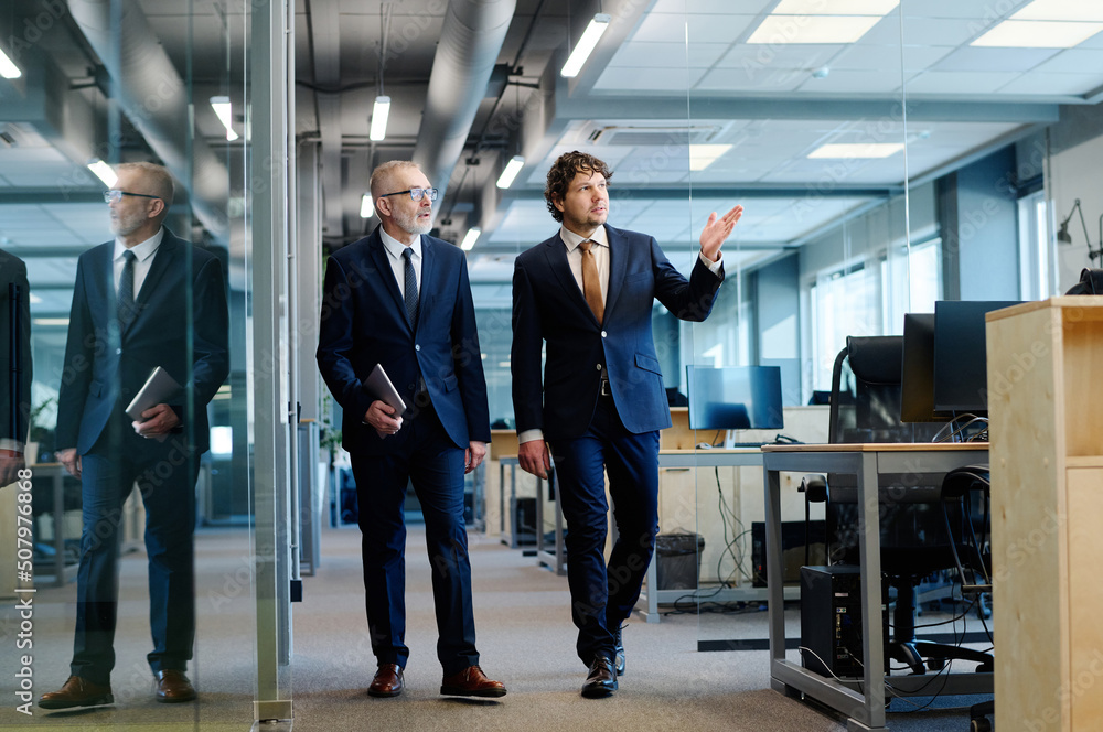 Young manager in suit showing the workplace to his mature colleague while they walking along office Young manager in suit showing the workplace to his mature colleague while they walking along office