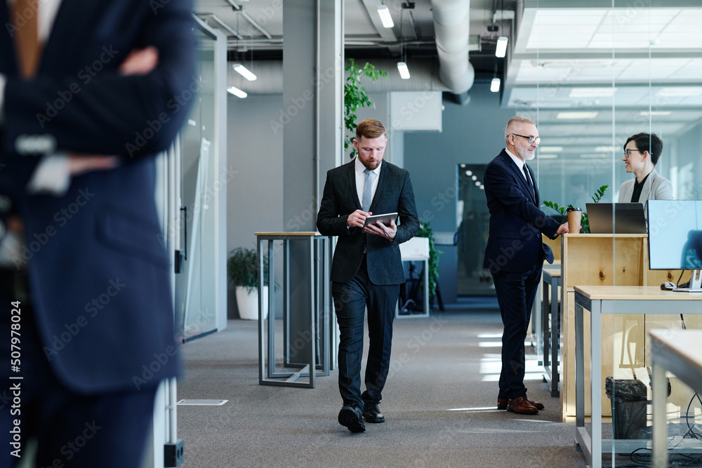 Obraz premium Young businessman using tablet pc walking along corridor with business people in formal clothes working in office