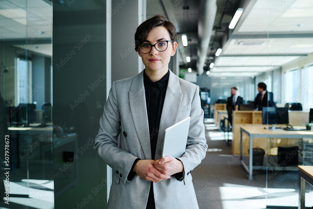 Portrait of beautiful young woman with short hair in formal suit holding digital tablet and looking at camera standing at office Portrait of beautiful young woman with short hair in formal suit holding digital tablet and looking at camera standing at office