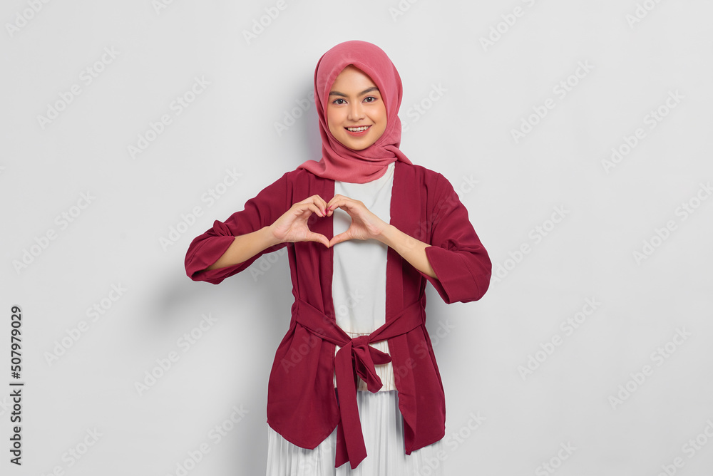 Smiling beautiful Asian woman in casual shirt making love gesture near chest isolated over white background