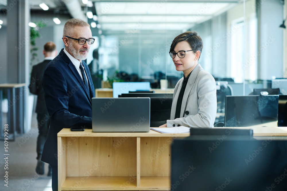 Young businesswoman in eyeglasses discussing online presentation on laptop with her mature colleague at office Young businesswoman in eyeglasses discussing online presentation on laptop with her mature colleague at office