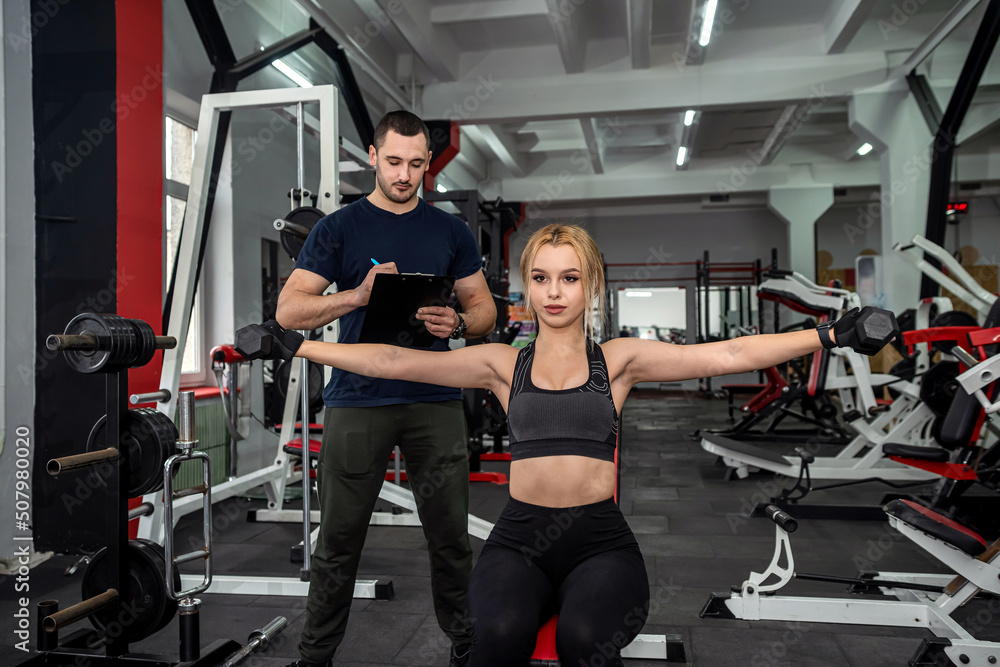 Personal male instructor helping woman doing exercise with heavy dumbbells