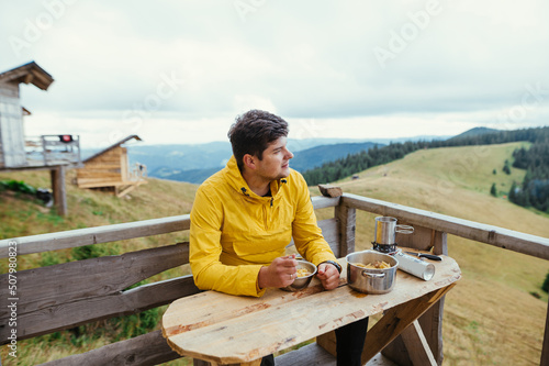 Handsome man in a yellow jacket eats in the mountains on the terrace of a country house and looks away with a smile on his face.