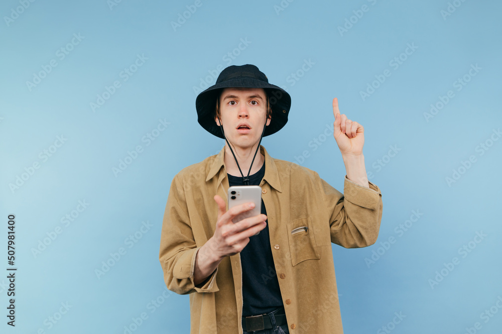 Surprised young man in panama and shirt stands on blue background with smartphone in hand and shows finger up with shocked face looking at camera. Surprised young man in panama and shirt stands on blue background with smartphone in hand and shows finger up with shocked face looking at camera.