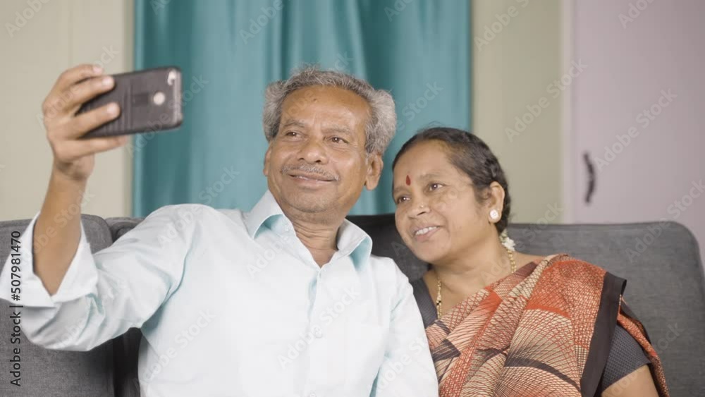 happy senior couple taking selfie on mobile phone while sitting on sofa at home - concept of family, togetherness and bonding.