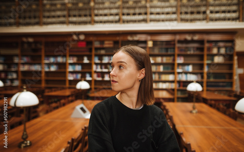 Portrait of a beautiful woman in dark clothes standing in the public library of the university campus and looking away.