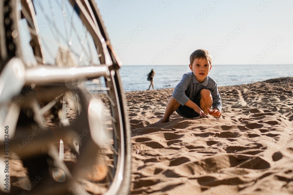 Cute little caucasian boy sitting on beach by father bicycle. Family weekend in countryside