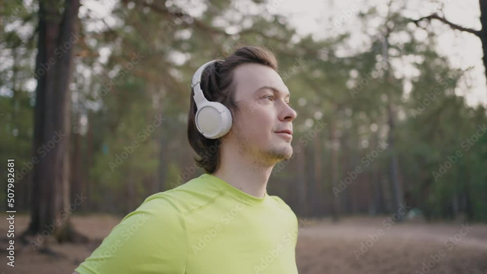 Runner in wireless headphones watches landscape during training break. Young man in pine forest looks at nature beauty listening to music Runner in wireless headphones watches landscape during training break. Young man in pine forest looks at nature beauty listening to music