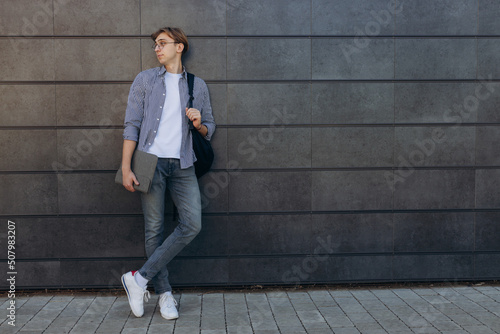 Happy student in glasses with laptop. Portrait of a young man with textbooks in his hands. Smiling boy with a backpack on dark wall. The concept of education. Copy space © Igor