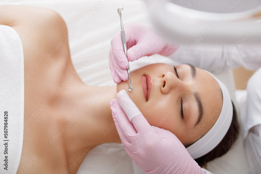 Young woman gets facial cleansing in a cosmetologist's office. Skin cleaning at beauty salon. Beautician squeezing pimples and blackheads from face.