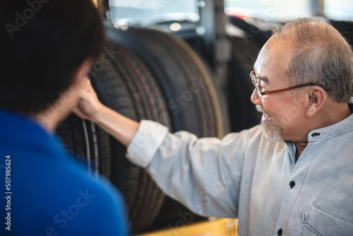 Smiling senior man with beard and spectacles checking car tires from rack in garage while getting assistance from young male mechanic