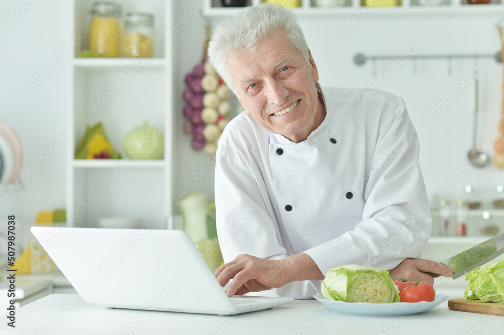 Elderly male chef cooking at table with laptop Elderly male chef cooking at table with laptop