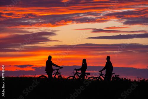 Image of sporty company friends on bicycles outdoors against sunset mountain. Silhouette of  three cyclist along sunset sky.