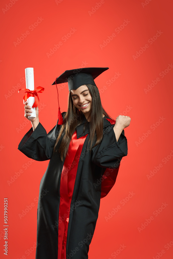 Front view of glad girl with bachelor, master degree holding diploma. Pretty female standing with closed eyes, showing hurray, smiling. Isolated on red studio background. Front view of glad girl with bachelor, master degree holding diploma. Pretty female standing with closed eyes, showing hurray, smiling. Isolated on red studio background.