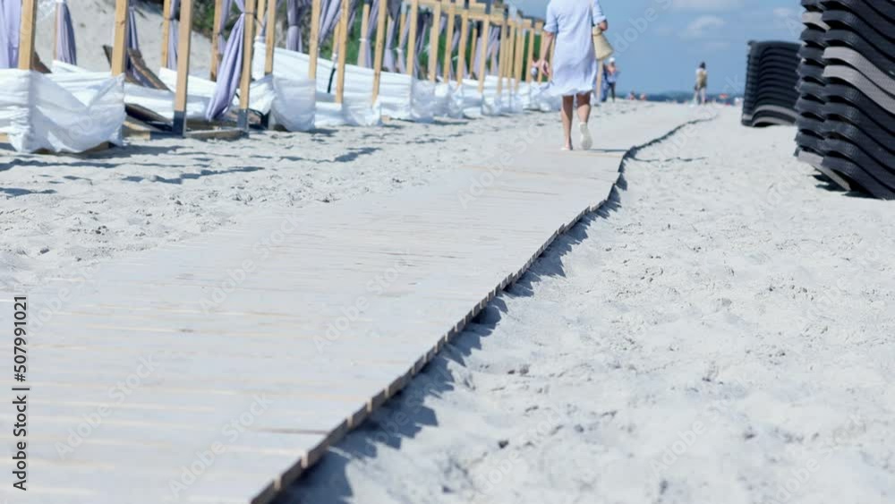 female barefoot legs walking wooden path along sandy beach sunny summer day. holiday season preparations deckchairs and chaise lounges standing on beach sand. hand holding shoes in hands. female barefoot legs walking wooden path along sandy beach sunny summer day. holiday season preparations deckchairs and chaise lounges standing on beach sand. hand holding shoes in hands.
