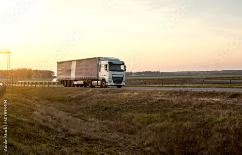 A semitrailer tractor with a tilt semitrailer transports cargo against the backdrop of an evening sunny sunset in summer. The concept of the logistics system and the business of cargo transportation