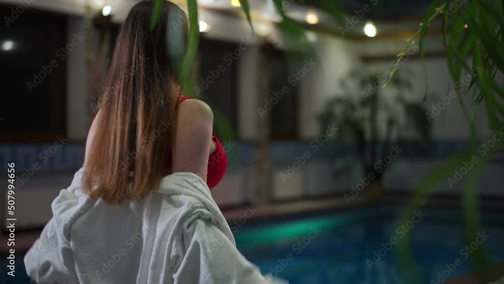 Seductive young Caucasian woman taking off white bathrobe walking to pool in red bikini. Young beautiful slim female tourist strolling in slow motion in hotel indoors. Tourism and femininity concept Seductive young Caucasian woman taking off white bathrobe walking to pool in red bikini. Young beautiful slim female tourist strolling in slow motion in hotel indoors. Tourism and femininity concept