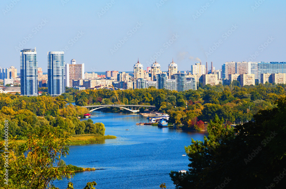 Obraz premium Scenic autumn landscape view of Kyiv. Nature landscape with autumn colors. Dnipro River and Hydropark against blue sky. Modern colorful residential skyscrapers in the background. Kyiv, Ukraine