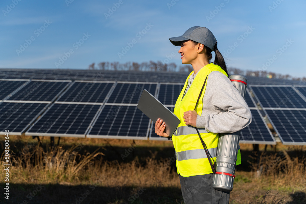 Side view of the smart lady engineer using electronic tablet monitoring solar panels and smiling happy. Sustainable energy, solar power field, ecology concept Side view of the smart lady engineer using electronic tablet monitoring solar panels and smiling happy. Sustainable energy, solar power field, ecology concept