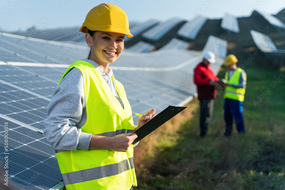 Obraz Beautiful female engineer technologist standing among solar panels and holding documents. Woman in protective helmet and uniform smiling at camera fototapeta, plakat