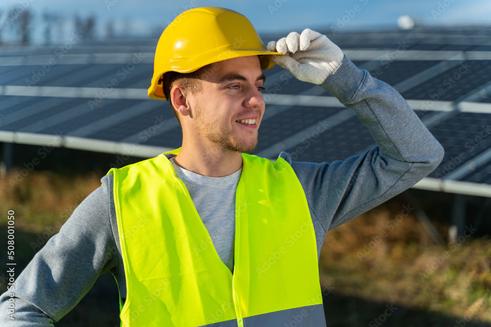 Male technologist in uniform with protective helmet. Adult man looking away while standing on field with solar panels Male technologist in uniform with protective helmet. Adult man looking away while standing on field with solar panels