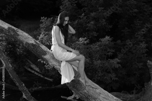 Fototapeta Naklejka Na Ścianę i Meble -  Beautiful long-haired young woman sitting on an old tree in the middle of the lake in forest in black and white